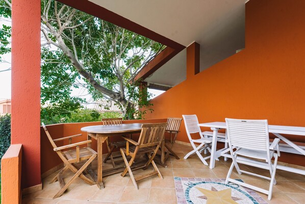 An outdoor patio with a wooden table and chairs, a tiled floor, and a view of trees and a building in the background.
