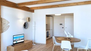 Une cuisine moderne avec des poutres en bois, un coin repas avec des chaises blanches et un téléviseur posé sur un meuble en bois.