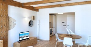 Une cuisine moderne avec des poutres en bois, un coin repas avec des chaises blanches et un téléviseur posé sur un meuble en bois.