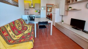 A compact living space with a sofa, a dining table, chairs, a TV, a ceiling fan, and a kitchen area with a refrigerator and microwave. - Luisa 5B - Cozy three rooms apartment (Lido di Pomposa)