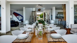 Un comedor con una mesa larga dispuesta para una comida, una cocina al fondo y una escalera que sube a otro nivel.