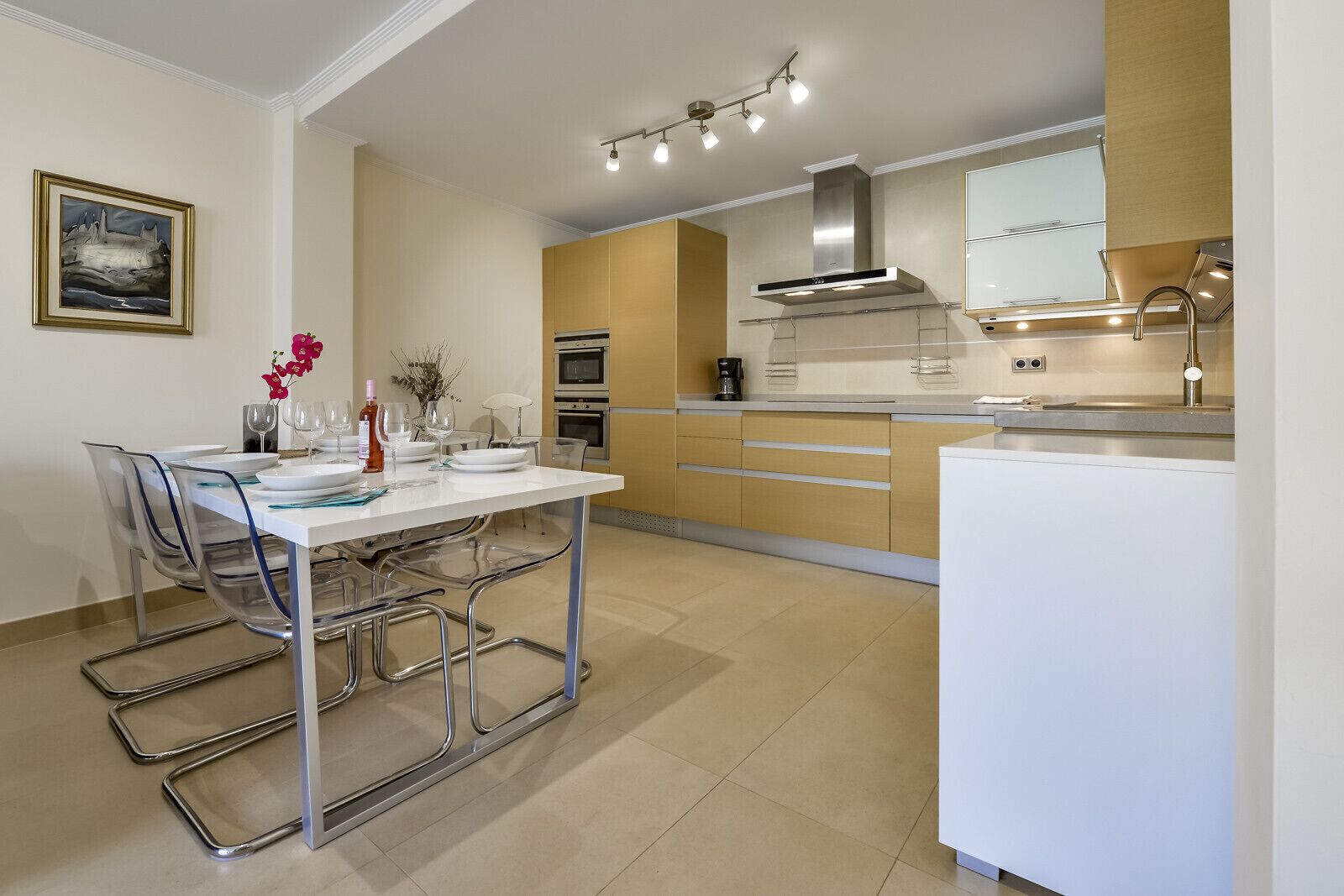 A modern kitchen with a dining area, featuring a white table, clear chairs, and a coffee machine.
