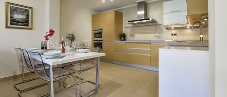 A modern kitchen with a dining area, featuring a white table, clear chairs, and a coffee machine.