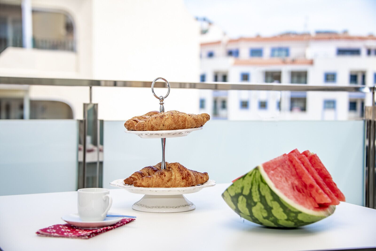 Une table surmontée d’un présentoir à trois niveaux, où sont disposés des croissants, une tasse et une tranche de pastèque, sur un balcon avec un immeuble en arrière-plan.