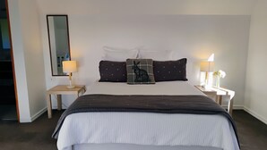 A neatly made bed with a dark brown headboard, white bedding, and two decorative pillows. There are bedside tables with lamps and a mirror on one side.