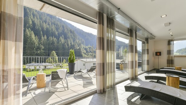 A balcony with outdoor seating, a view of a forested mountain, and a modern interior with large windows and curtains. - Studio in der Alpine Lodge Arlberg (Klösterle am Arlberg)