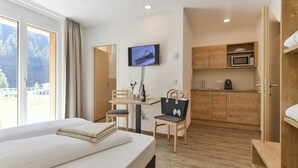 A hotel room with a bed, a desk with a chair, a television, a kitchen area with wooden cabinets, and a view of the outdoors. - Studio in der Alpine Lodge Arlberg (Klösterle am Arlberg)