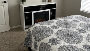 Een slaapkamer met een open haard, een ronde spiegel, een witte console tafel met decoratieve objecten, en een bed met een bekleed patroon.