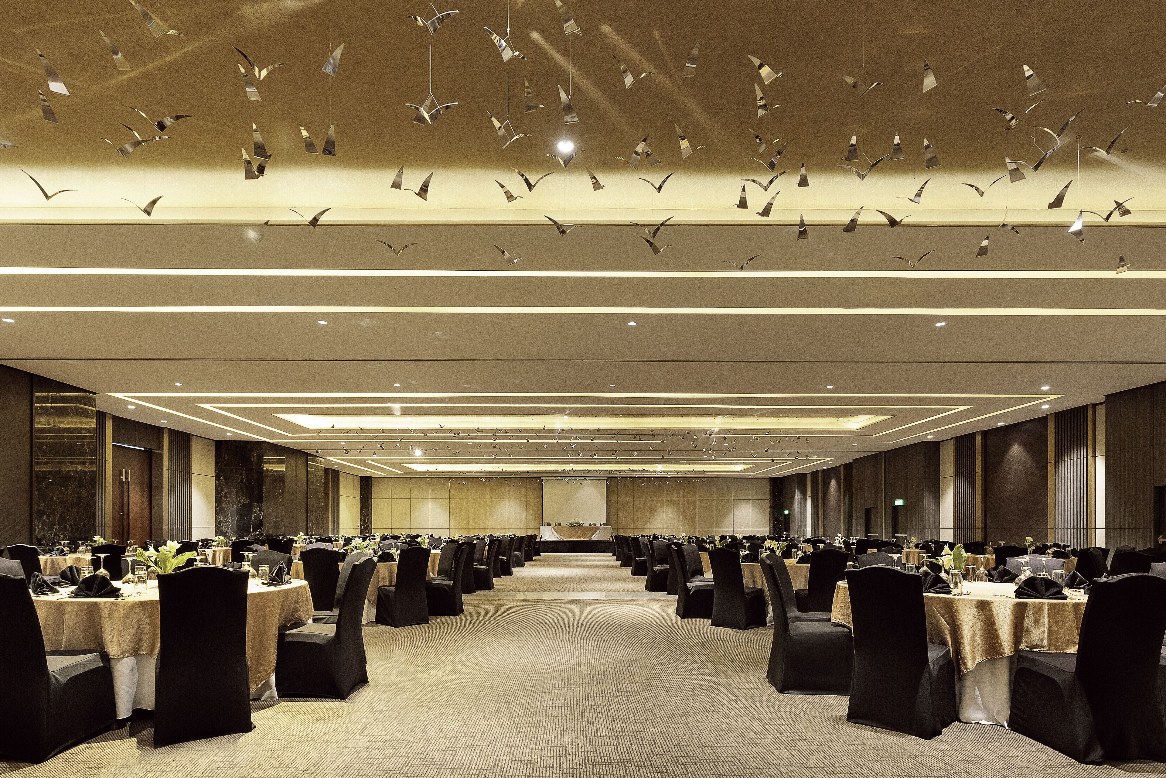 Un amplio salón de banquetes con filas de mesas y sillas, un escenario al frente y un techo con lámparas colgantes.