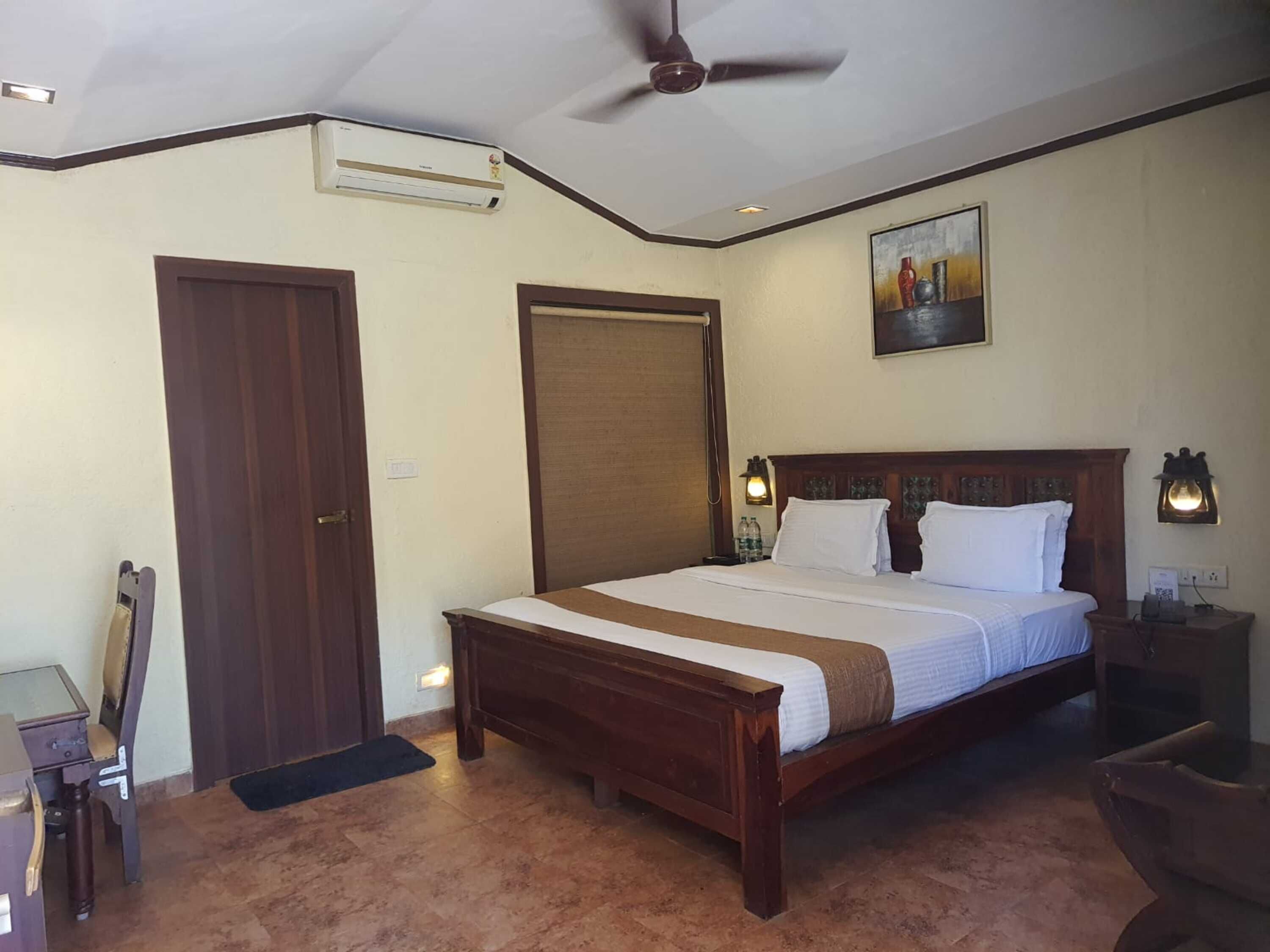 A hotel room with a large bed, a ceiling fan, an air conditioning unit, a door, a painting on the wall, and a nightstand with a lamp.