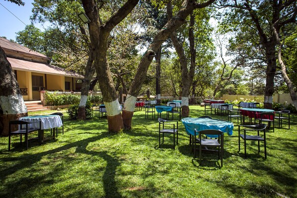 Un área de estar al aire libre con mesas y sillas, rodeada de árboles y un edificio con techo inclinado.