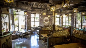 A restaurant with high ceilings, exposed brick walls, and ornate metal railings. Tables and chairs are arranged for dining, and large windows allow natural light to enter. - Adamo The Village (Karjat)