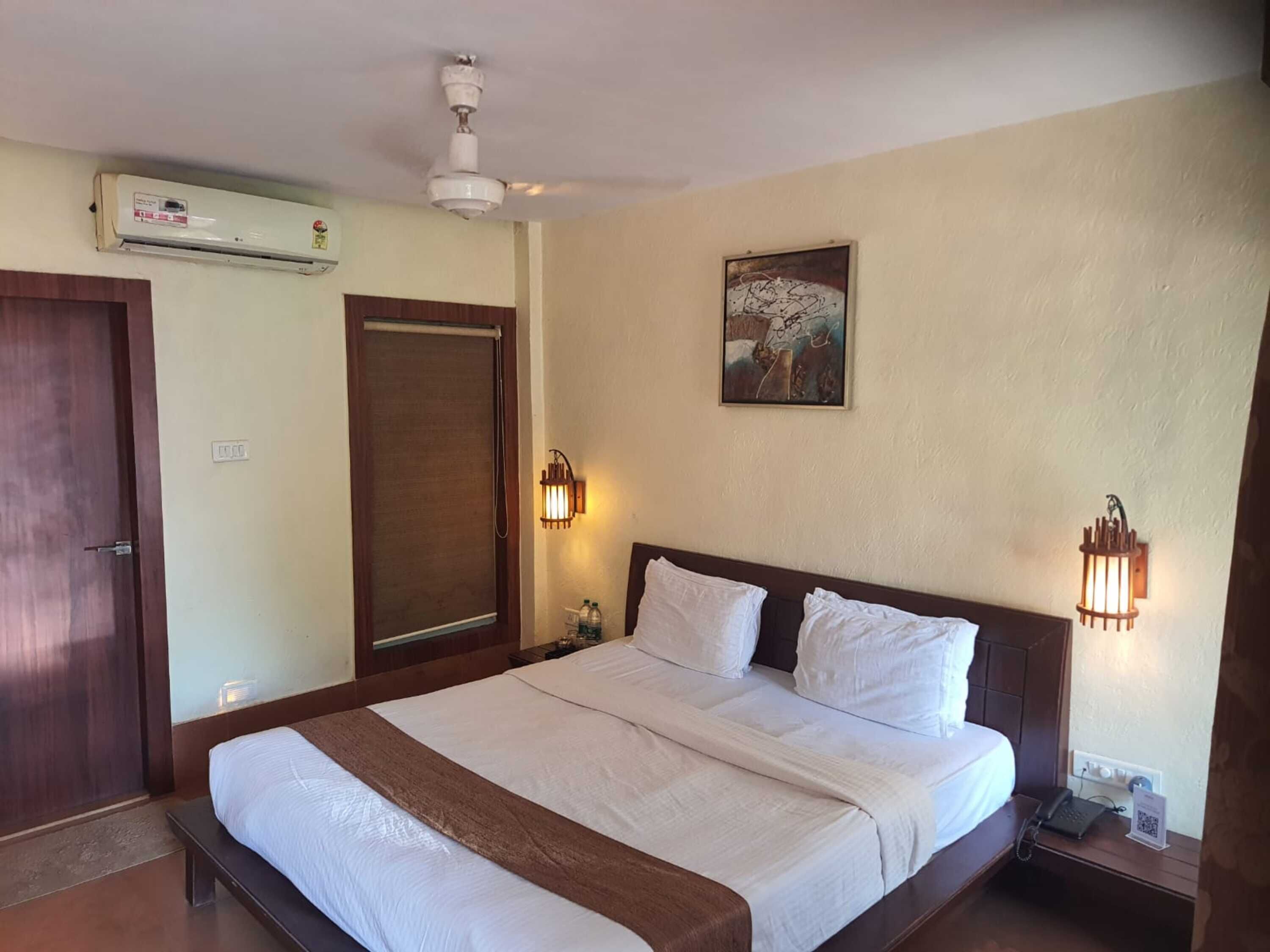 A hotel room with a large bed, a ceiling fan, an air conditioning unit, a door, a wall-mounted artwork, and two wall sconces.