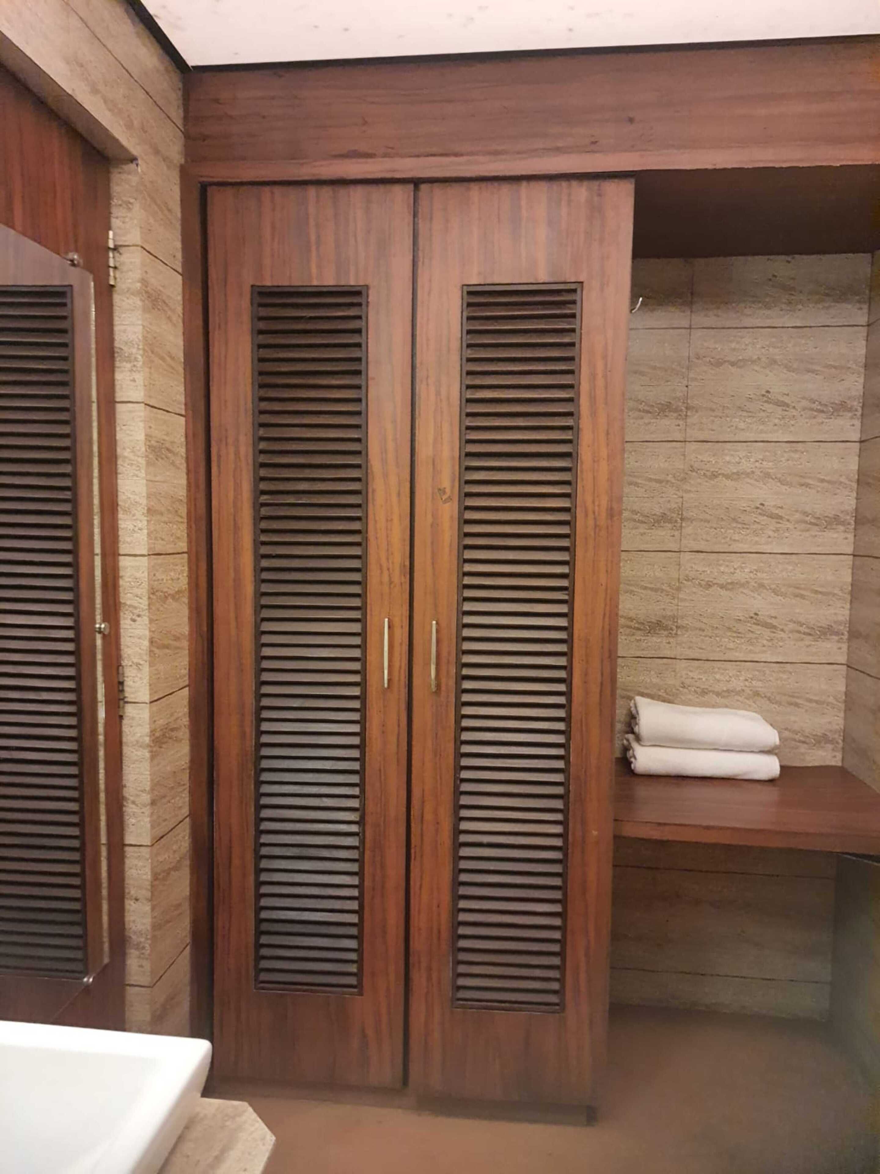 A corner bedroom with a wooden wardrobe, a small shelf with folded towels, and a stone-textured wall.