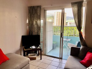 Una sala de estar con un sofá, un televisor y un ventanal corredera que da a un balcón con vistas a edificios y zonas verdes.