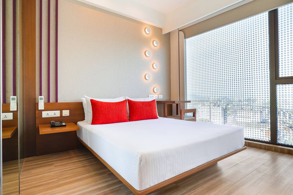 A modern hotel room with a large bed, wooden headboard, red pillows, bedside tables, and a large window with a city view.