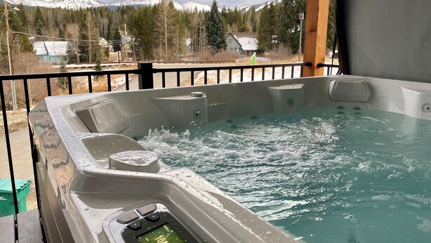 Un jacuzzi con pantalla digital, rodeado por una cubierta de madera y una barandilla metálica, con vistas a un paisaje montañoso nevado.