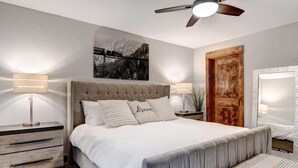 Un dormitorio con cabecera capitonada, una cama grande con ropa de cama blanca, dos mesitas de noche con lámparas, un banco al pie de la cama, una puerta de madera y un cuadro enmarcado en la pared.