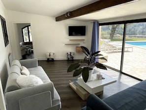 A modern living room with a grey sofa, a coffee table with a plant, a flat-screen TV mounted on the wall, and a view of a swimming pool through large windows. - Villa-Monti, Menton, Côte d'Azur (Menton)
