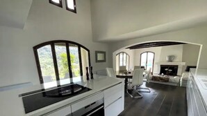 A modern kitchen with a central island, white cabinetry, and a black countertop. There is a dining area with a white table and chairs, and a fireplace in the background. - Villa Monti: Luxury Villa, Large Pool, Air Con, Amazing Views (Menton)