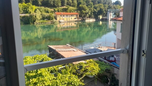 Blick auf einen Fluss mit einem Gebäude am gegenüberliegenden Ufer, einem Balkon mit Geländer und Grünpflanzen im Vordergrund.