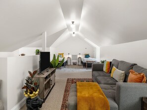 A modern loft bedroom with a sloped ceiling, a large gray sofa, a small bed, and a television on a wooden stand.