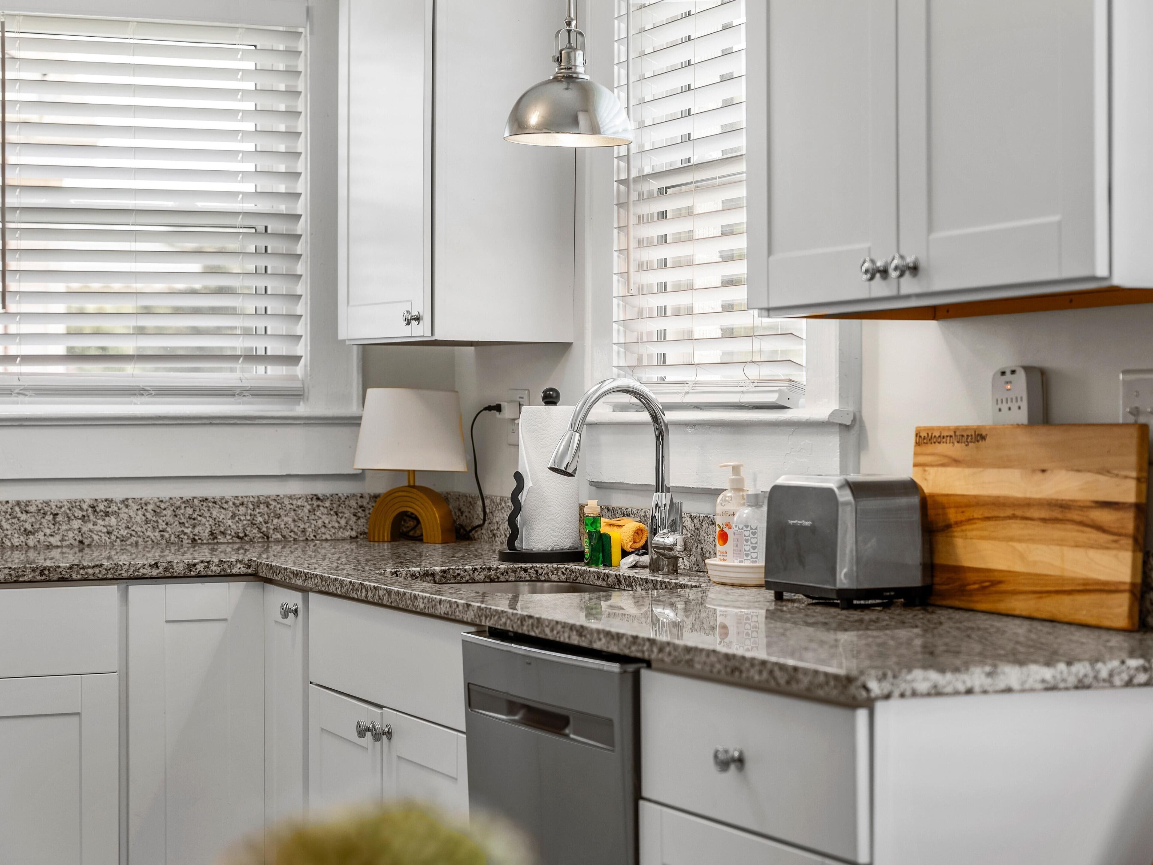A kitchen with white cabinets, a granite countertop, a sink, a dish rack, a lamp, and a cutting board.