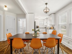 A dining area with a wooden table and orange chairs, a kitchen in the background, and a ceiling light fixture. - Jungalow Retreat near Swamp Rabbit Trail! (Greenville)