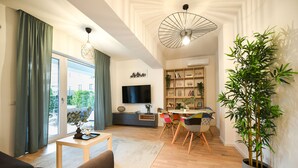 Ein modernes Wohnzimmer mit einer Couch, einem Couchtisch und einem Essbereich mit Stühlen. Ein Fernseher, ein Bücherregal und eine große Pflanze sind ebenfalls vorhanden.