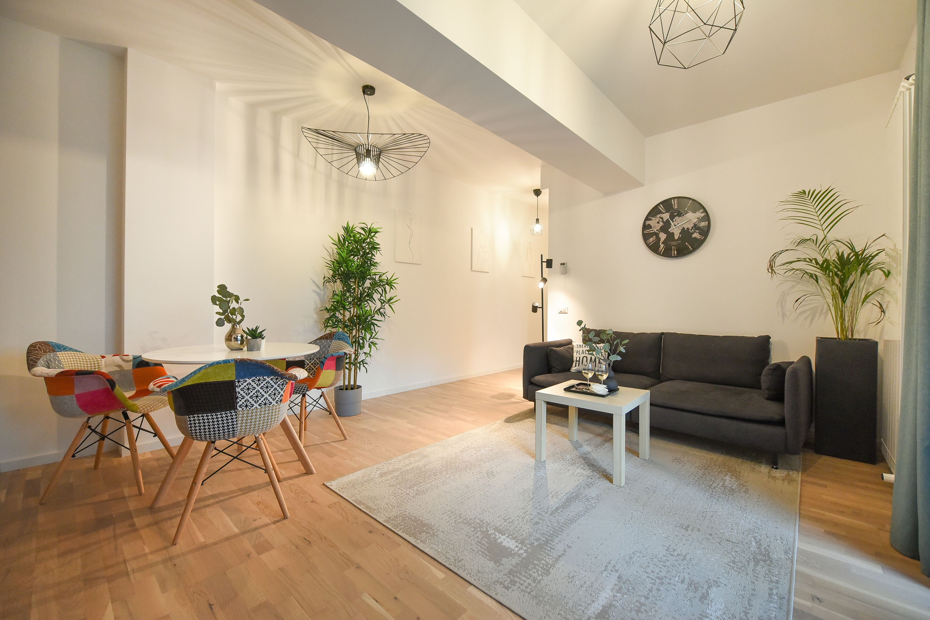 A modern living room with a dining area, featuring a sofa, a coffee table, a dining table with colorful chairs, and potted plants.