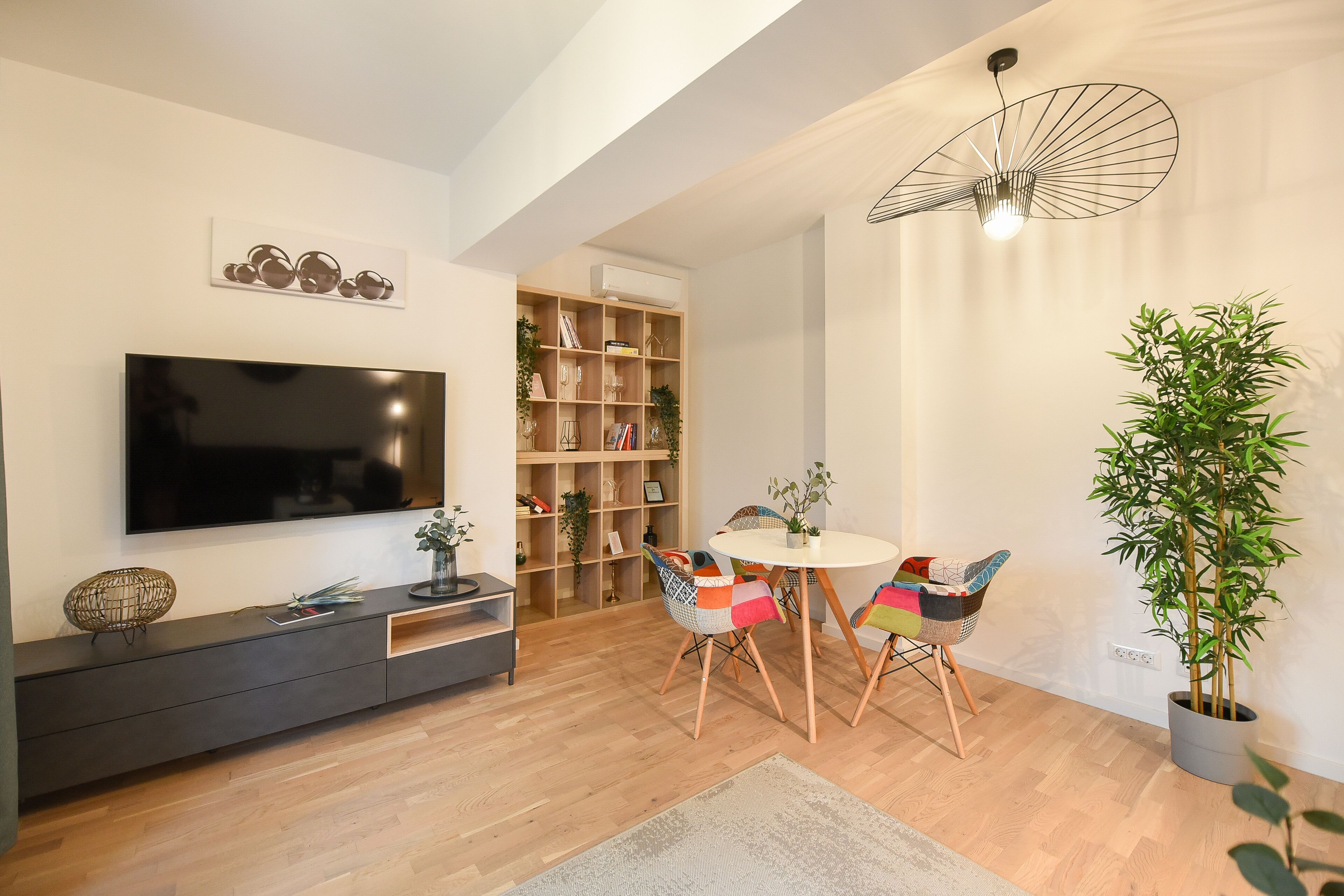 Un salon moderne avec un téléviseur à écran plat, une bibliothèque, une table à manger avec des chaises colorées et une plante en pot.