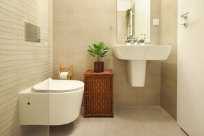 A modern bathroom with a white toilet, a wicker side table with a plant, a white sink, and a mirror above the sink. - Vacation Feel (Bucharest)