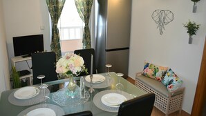 Un coin repas avec une table en verre dressée pour quatre personnes, un réfrigérateur, un téléviseur à écran plat et un canapé agrémenté d’un coussin à motifs floraux.