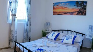 Un lit bien fait, décoré sur le thème de la marine, un tableau représentant un coucher de soleil et une fenêtre avec des rideaux.