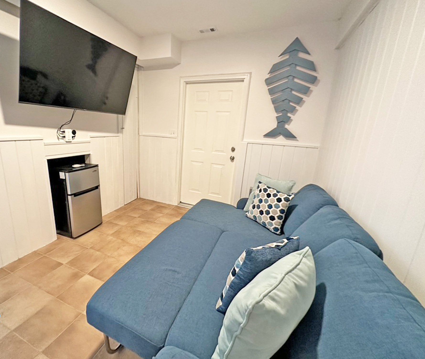 Ein kleines Zimmer mit Bett, Wandfernseher, kleinem Kühlschrank und einer Tür.