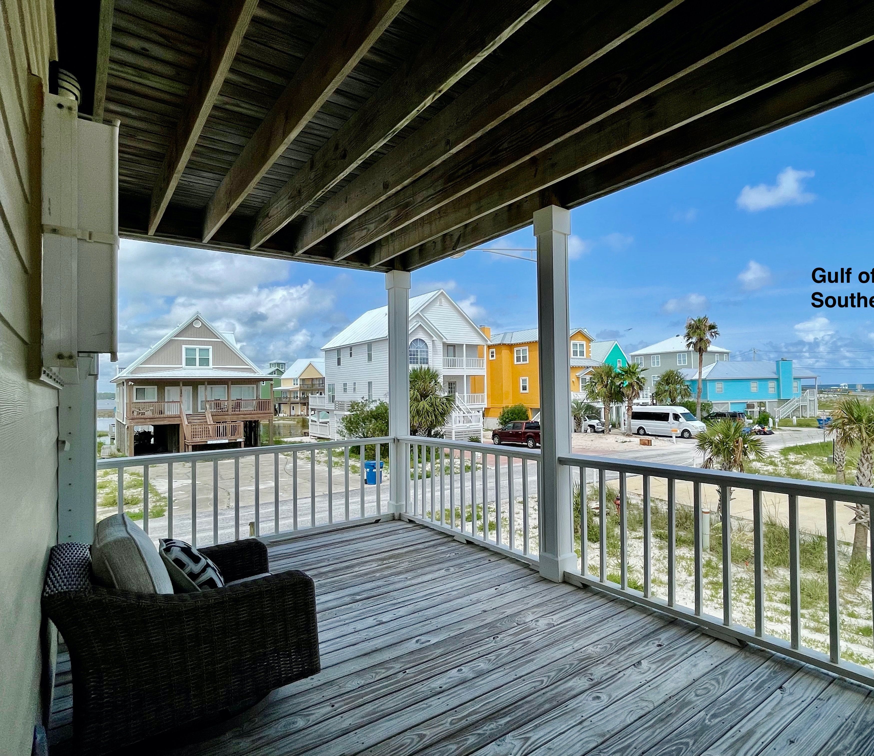 A covered porch with a view of the Gulf of Mexico, featuring a wooden deck, wicker furniture, and a railing.