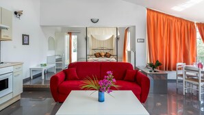 Une chambre moderne avec un canapé rouge, une table basse blanche ornée d’un vase de fleurs, un lit à baldaquin et un coin repas avec des rideaux orange.