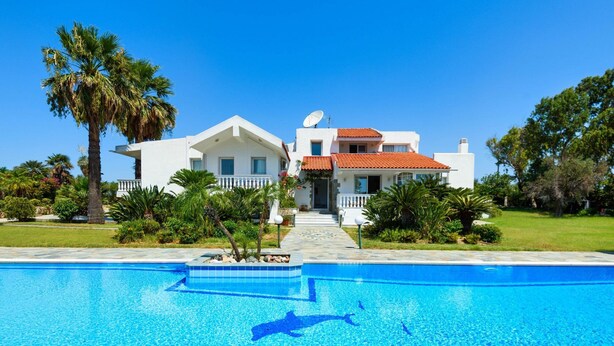 Une grande maison avec une piscine, des palmiers et un ciel d’un bleu éclatant.