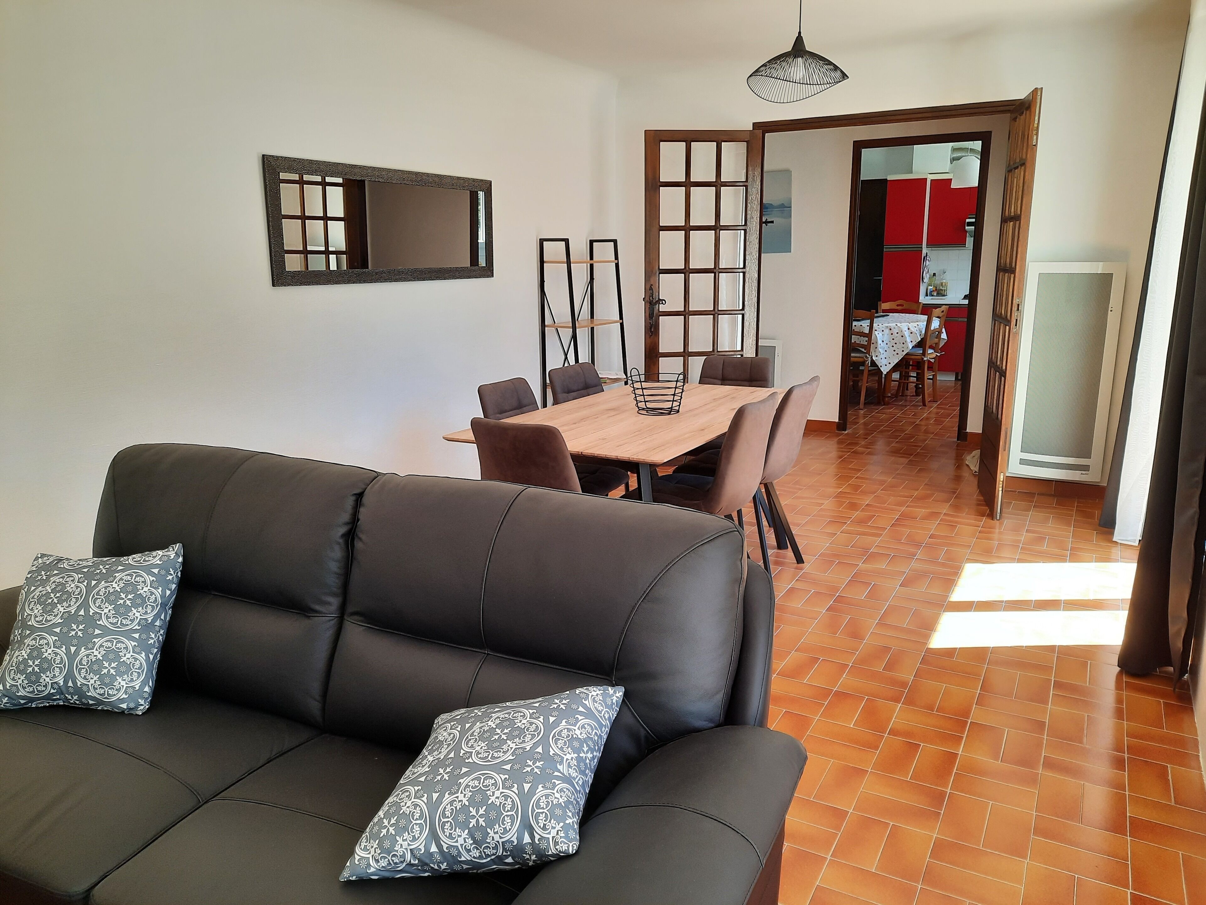 Una sala de estar con un sofá de cuero oscuro, cojines estampados, una mesa de comedor de madera y sillas. Un espejo grande cuelga en la pared, y una puerta corrediza da acceso a una zona de cocina.