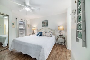 Un dormitorio con una cama grande, un ventilador de techo, dos mesitas de noche con lámparas, un espejo y una puerta que da a otra habitación.