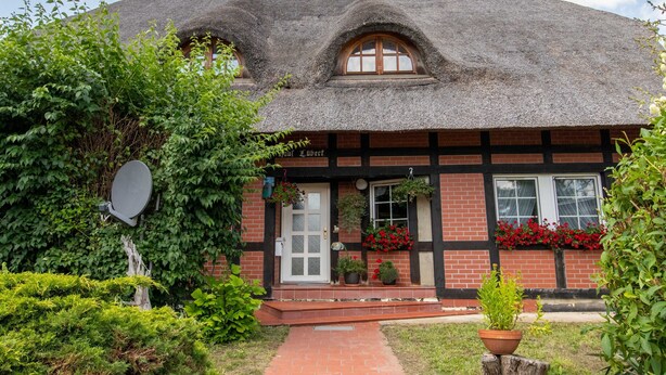 En traditionel bygning med stråtækt tag, murstensfacade og en lille veranda med plantekrukker.