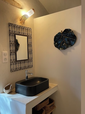 Un baño con un lavabo tipo cuenca negro, un espejo decorativo con marco de hierro forjado y una lámpara de pared.
