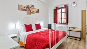 A bedroom with a white metal bed frame, a red and white bedspread, a bedside table with a lamp, a wall-mounted artwork, a window with red curtains, and a small bedside table with a plant.