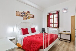 Une chambre avec un lit en métal blanc, une couvre-lit rouge et blanche, une table de chevet avec une lampe, une œuvre d’art accrochée au mur, une fenêtre avec des rideaux rouges et une petite table de chevet ornée d’une plante.