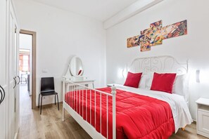 A bedroom with a white bed, red bedding, a desk with a chair, and a wall with colorful artwork. - GuestHost - Comfortable 35sqm apartment that can accommodate up to 4 people. Located on the second floor, without a lift, of a building 3 minutes walk away from Uffizi Gallery and Piazza della Signoria, the accommodation is the best solution to visit the (Firenze)