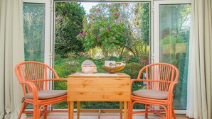 A wooden table with two orange wicker chairs, a bowl, and a book on it, set on a patio with a view of a garden with pink flowers and green trees. - Garden Pool House by Liiiving (Porto)