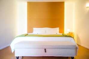 A bed with white bedding and a green throw, a bedside lamp, and a plain wall with a wooden panel above the bed.