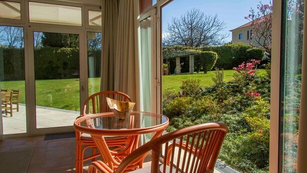 A glass-enclosed patio with a round table and orange chairs, overlooking a well-manicured garden and a house. - Garden Pool House by Liiiving (Porto)