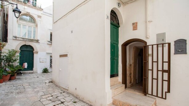 A narrow cobblestone street with a white building featuring a green door and a wooden gate. - GuestHost - Lovely apartment located in the heart of Monopoli, in the historic center, in a stone building of which it retains suggestive evidences in the internal architecture. The flat can comfortably accommodate two people and is located in an enviabl (Monopoli)