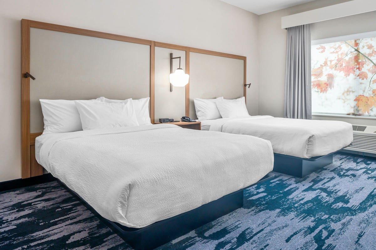 A hotel room with two beds, a nightstand, a lamp, and a large window with a view of autumn leaves.